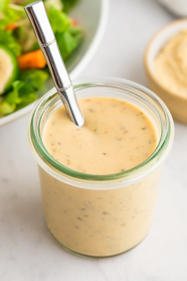 How to make the BEST curried hummus dressing plus a delicious rainbow kaleidoscipe salad! A jar of easy hummus dressing. A silver spoon is poking out of the top.