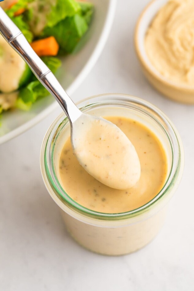 How to make the BEST curried hummus dressing plus a delicious rainbow kaleidoscope salad! A jar of easy hummus dressing with a spoon ladling some dressing out.