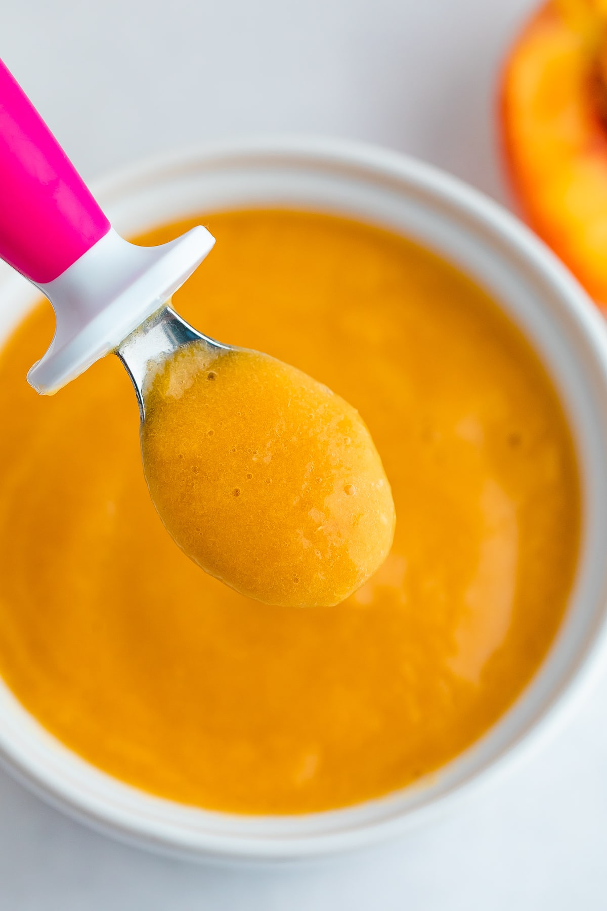 Up close photo of peach puree on a pink baby spoon hovering over a white bowl with peach baby food.
