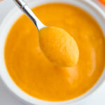 Peach puree on a small sliver spoon being held over a white bowl with the puree.
