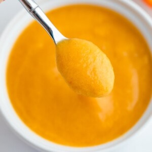 Peach puree on a small sliver spoon being held over a white bowl with the puree.
