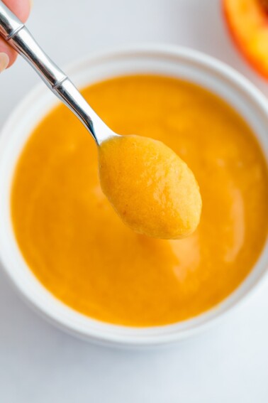 Peach puree on a small sliver spoon being held over a white bowl with the puree.