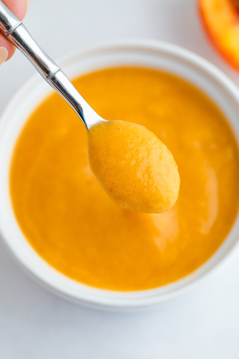 Peach puree on a small sliver spoon being held over a white bowl with the puree.