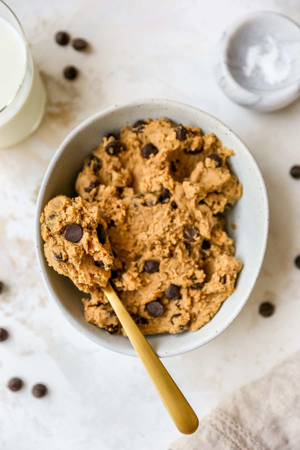 PIN THIS--> Protein Cookie Dough Edible protein cookie dough in a white speckled bowl with chocolate chips on top and gold spoon on the left.