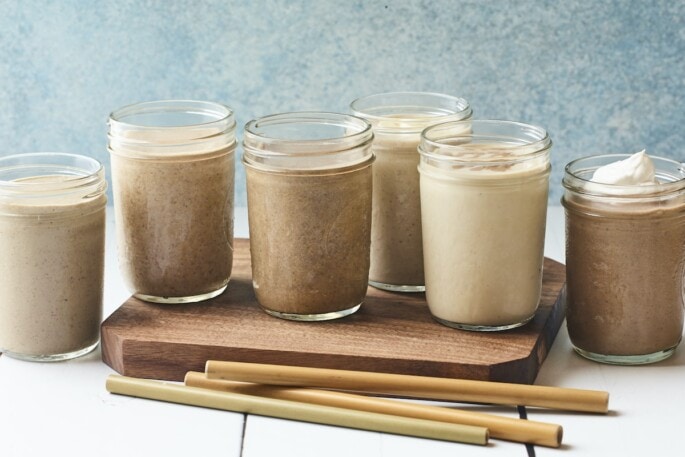 6 protein shakes in Mason jars on a wood board. Bamboo straws are on the table.
