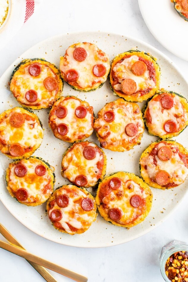 Mini Zucchini Hummus Pizza Bites -- Satisfy your pizza craving with these healthy mini zucchini pizza bites. Raw zucchini serves as the crust while hummus and fresh veggie toppings give the bites that pizza flavor you crave. Zucchini pepperoni pizza bites on a plate.