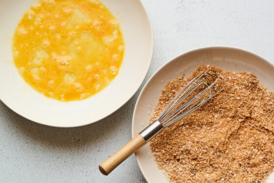 One bowl has eggs that have been whisked and the second bowl has a mixture of dry ingredients with a whisk resting in the bowl.