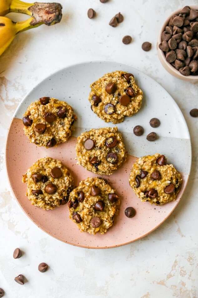 An overhead photo of 6 cookies on a plate with chocolate chips sprinkled around. You can also see a banana and cup of chocolate chips in the top corners.