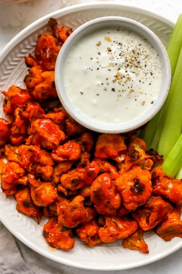 Looking for a healthy game day snack? Try these buffalo cauliflower wings with a creamy cashew dressing. They’re absolutely delicious and easy to whip up. Vegan + gluten-free. Plate of buffalo cauliflower wings served with vegan cashew ranch and celery sticks.