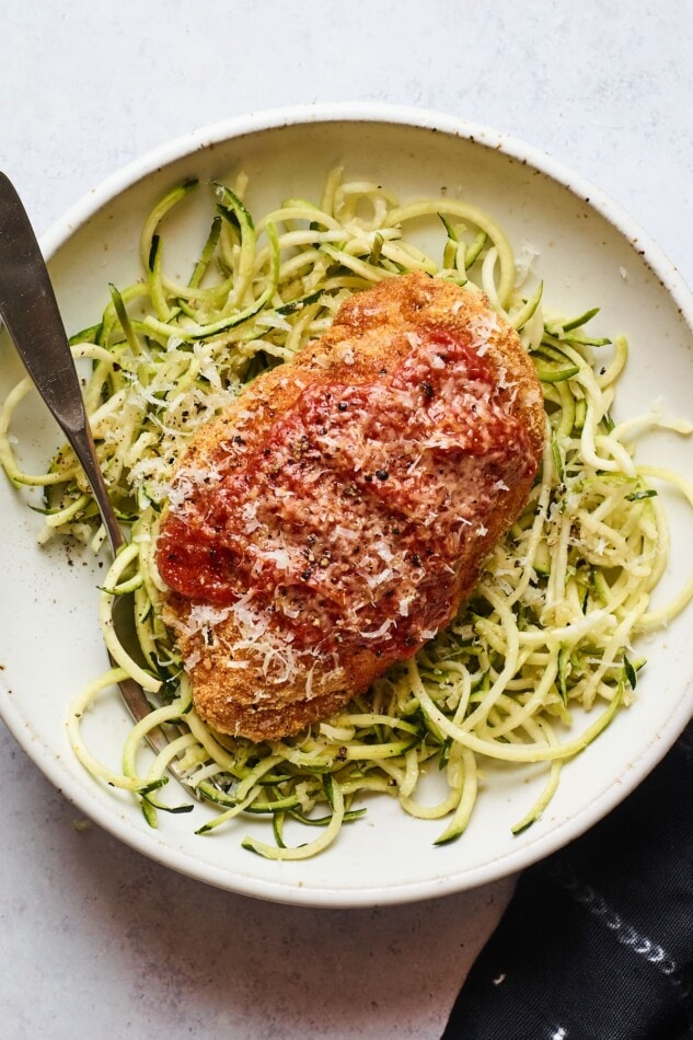 An overhead photo of a bowl zucchini noodles with a chicken parm breast resting on top. A fork rests inside the bowl.