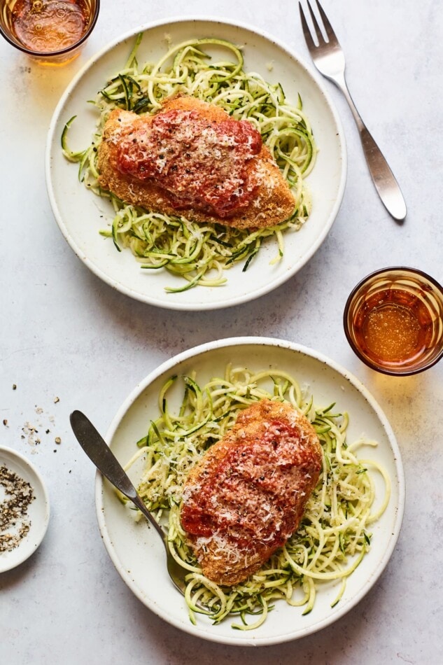 Overhead shot of a healthy dinner dish with chicken parmesan and broccoli. Both topped with a little cheese. An overhead photo of two bowls of zucchini noodles with a chicken breast resting on top of each.