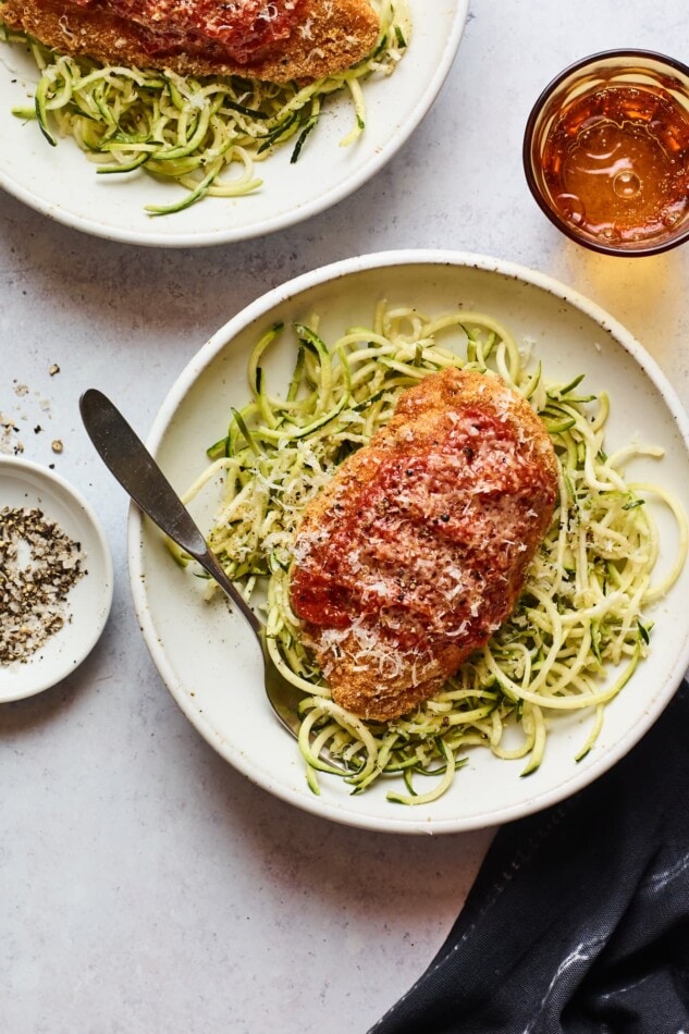 Overhead photo of a bowl of zucchini noodles with a chicken parm breast resting on top. There is a second bowl partially in the frame.