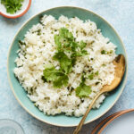 A serving of cilantro white rice topped with extra fresh cilantro leaves and a metal spoon to the side.