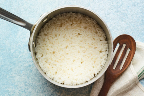 Cooked white rice in a stainless steel pot with a slated spoon off to the side.