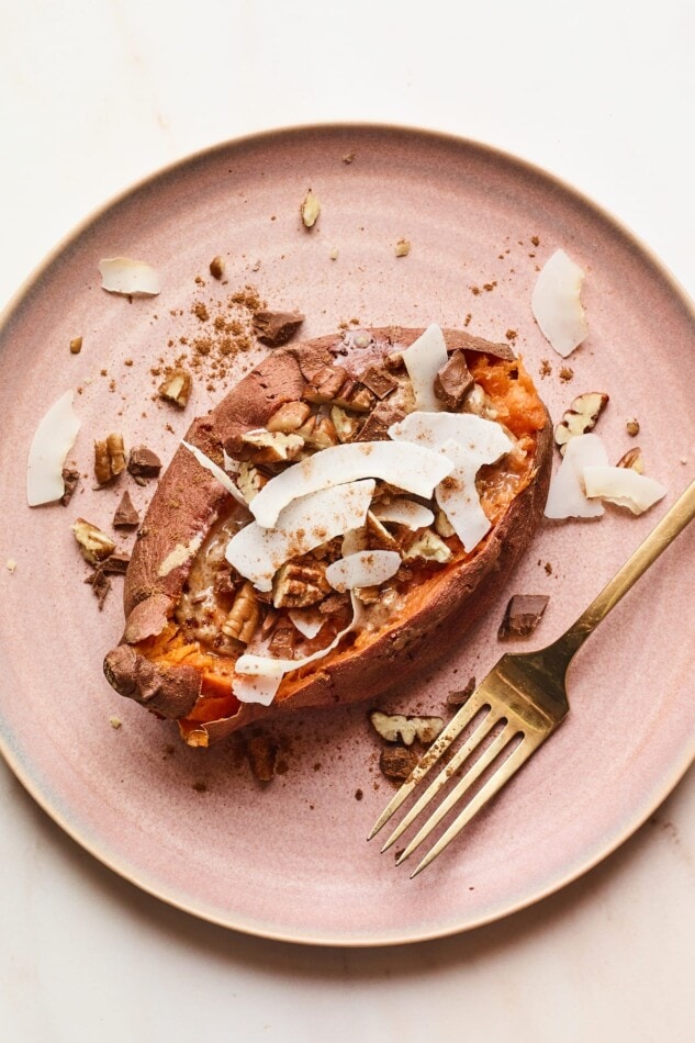 Turn nutrient-rich sweet potatoes into a satisfyingly sweet dessert! Just bake and stuff with your favorite toppings. This particular dessert baked sweet potato is loaded with toasted coconut, coconut butter, cinnamon, almond butter, pecans and dark chocolate. Vegan, gluten-free and paleo-friendly. An overhead photo of a dessert sweet potato with topping sprinkled around. A gold fork rests on the plate next to the potato.