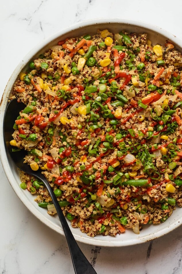 An overhead photo of a bowl of quinoa fried rice. Sriracha has been drizzled on top and a black serving spoon rests inside the bowl.