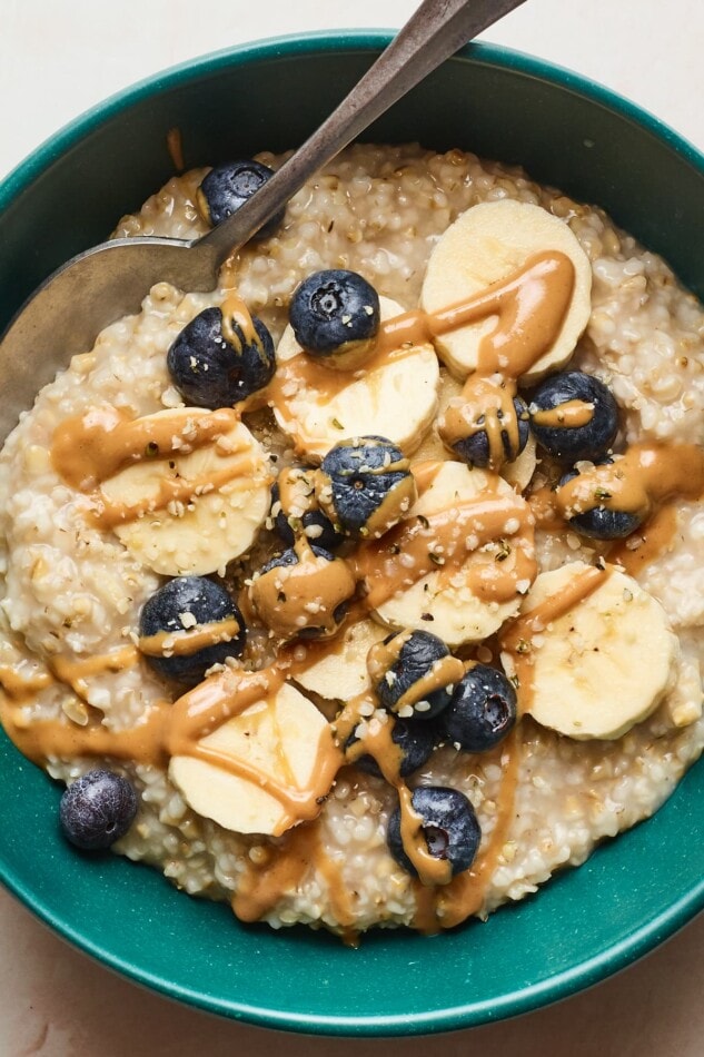 A close up photo of steel cut oats in a bowl with blueberries, bananas, and almond butter drizzled on top. A spoon rests in the bowl.