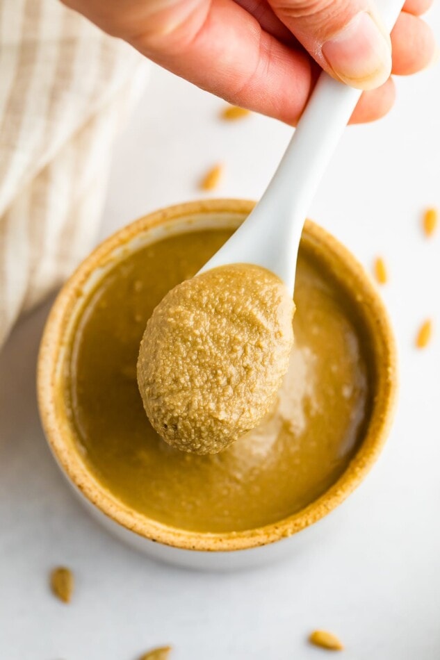 Make sunbutter at home with this simple and cost-effective recipe! Roasted sunflower seeds, maple syrup and vanilla make for a delicious sunflower seed spread that's great for those with nut-allergies and safe for school. Overhead photo of a hand spooning out some sunflower seed butter from open bowl.