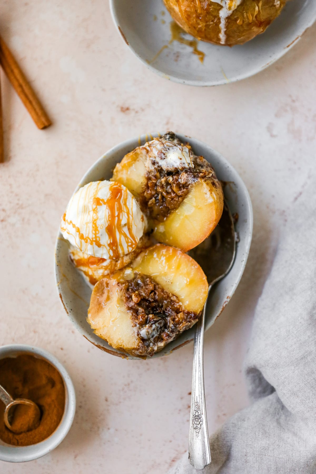 An overhead photo of a baked apple sliced in half with a scoop of ice cream and a caramel drizzle in a shallow bowl with a silver spoon.
