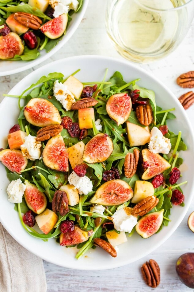 An overhead photo of a bowl of fig salad with a white wine glass next to it.