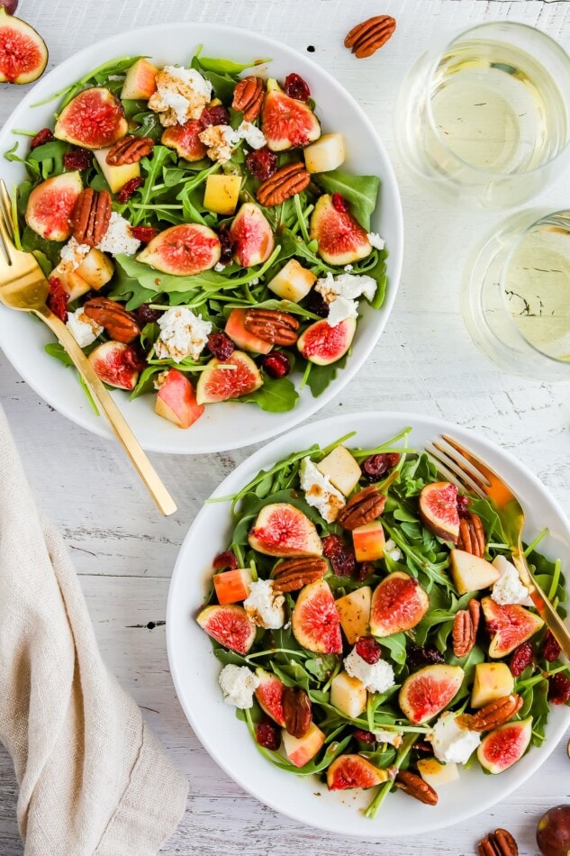 An overhead photo of two bowls of fig salad next to two glasses of white wine. A gold fork rests inside each bowl.