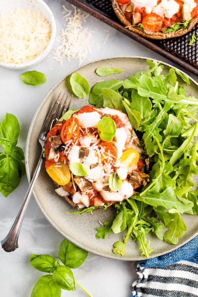 Overhead photo of a portobello mushroom pizza plated with arugula. A ramekin of cheese is in the top left corner and a silver fork rests on the plate.