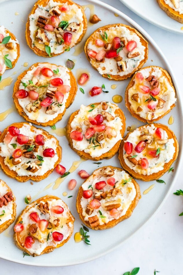 Overhead photo of a plate full of sweet potato bites topped with pomegranate arils, pecans, fresh thyme and drizzled with honey.