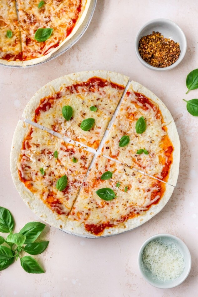 An overhead photo of a tortilla pizza with basil, cheese, and red pepper flakes in ramekins around the pizza.