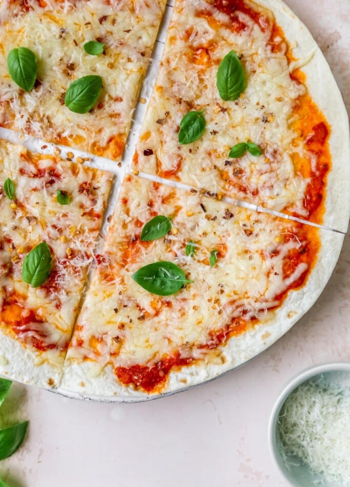 A close up overhead photo of a tortilla pizza. A ramekin of cheese is in the bottom right corner of the image.