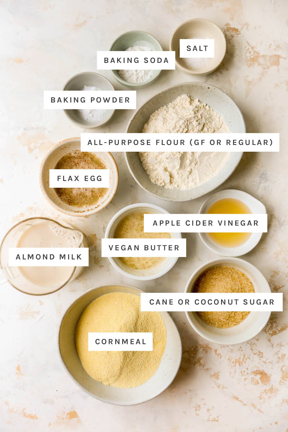 Ingredients measured out in little bowls to make vegan cornbread: salt, baking soda, baking powder, flour, flax egg, vinegar, vegan butter, almond milk, sugar and cornmeal.