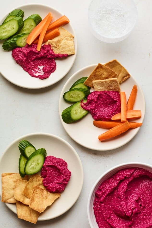 Beet hummus is bright pink in color, subtly sweet and full of flavor. Serve with veggies for a show-stopping party appetizer or use as a spread, dip or garnish. 3 plates with a scoop of beet hummus and various vegetables for dipping. A bowl of beet hummus is in the bottom right corner.