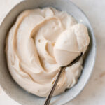 A small bowl of a cream cheese frosting with a metal spoon holding a spoonful of the frosting.
