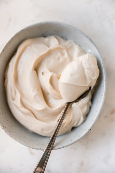 A small bowl of a cream cheese frosting with a metal spoon holding a spoonful of the frosting.