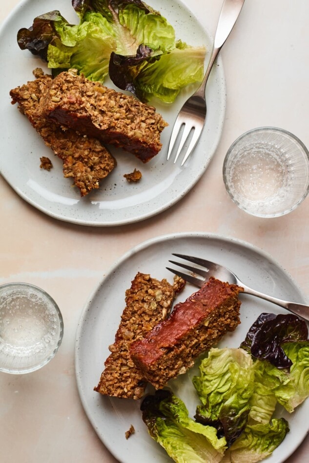 Save the recipe for this Lentil Loaf with a Maple Glaze! Two plates with two slices of lentil loaf and salad greens. Additionally, there are two forks and two glasses of water.