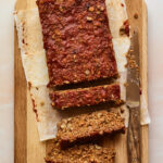 A vegan lentil loaf on a wooden cutting board with a knife nearby and three slices from the loaf are cut.