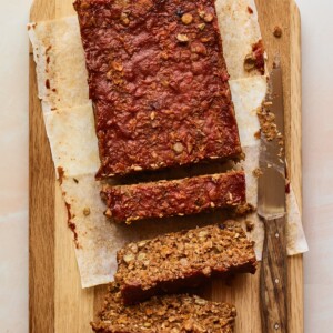 A vegan lentil loaf on a wooden cutting board with a knife nearby and three slices from the loaf are cut.