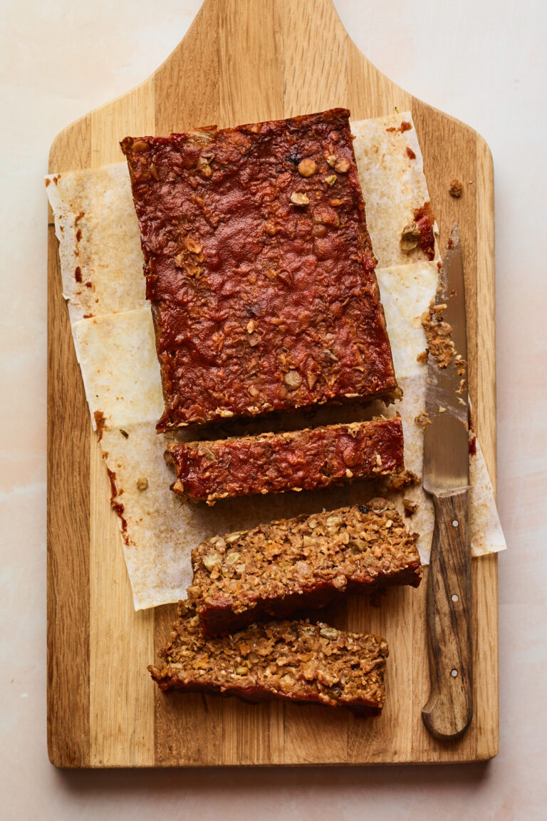 A vegan lentil loaf on a wooden cutting board with a knife nearby and three slices from the loaf are cut.