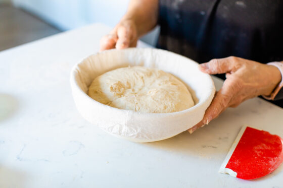 A woman is picking up a banneton basked with a ball of dough inside of it.