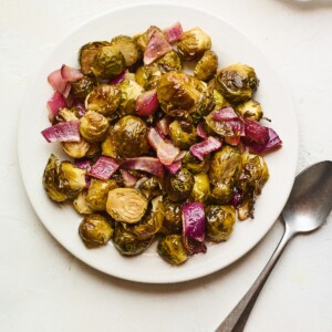A large serving of roasted brussels sprouts and red onion on a white plate with a metal spoon to the side.