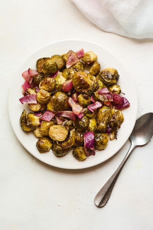 Roasted brussels sprouts and red onion in a white bowl with a gold spoon on the right. A serving of roasted brussels sprouts with red onion on a plate.