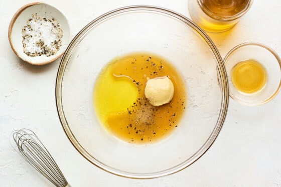 A glass bowl with vinegar, oil, maple syrup, mustard, salt and pepper ready to be whisked together. A whisk is laying next to the bowl.