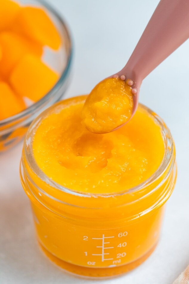 Butternut squash puree in a small baby food storage jar with a baby food spoon scooping some out.