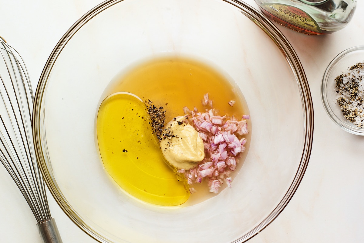 A glass mixing bowl containing vinegar, olive oil, maple syrup, minced shallots, dijon mustard, sea salt and pepper.