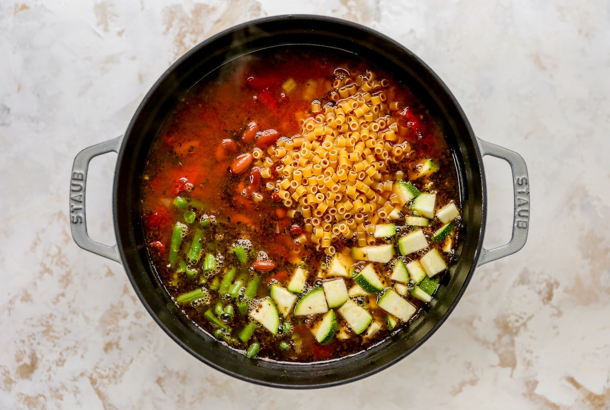 Additional minestrone soup ingredients added to a large pot, including green beans, zucchini, kidney beans and pasta.