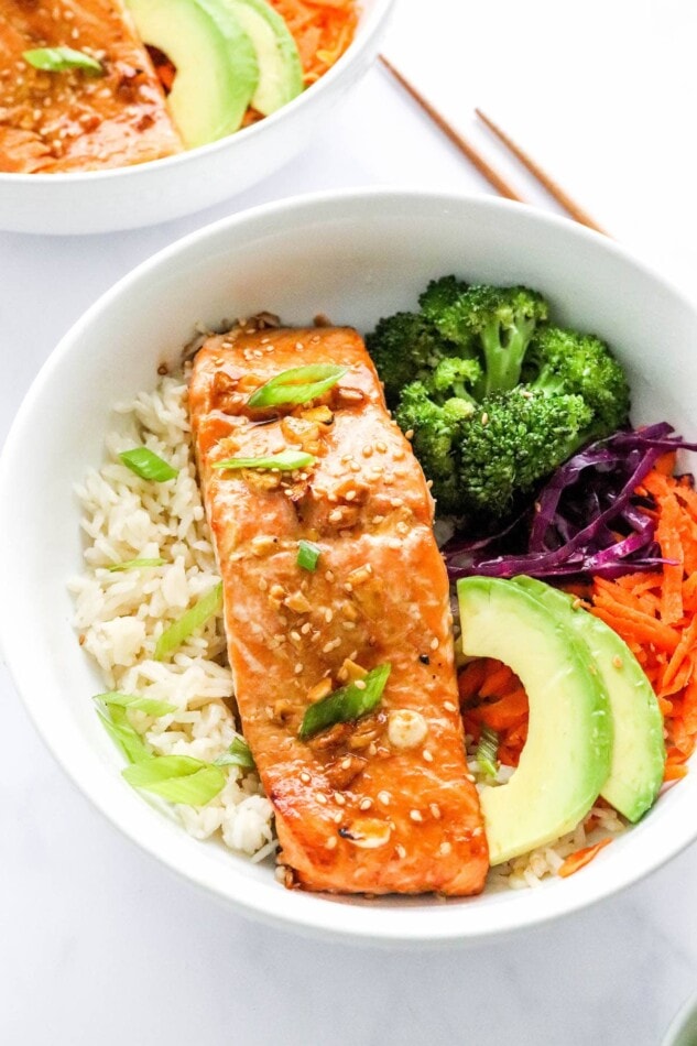These Teriyaki Salmon Bowls come together quickly for a healthy and delicious weeknight meal. Bake the salmon and broccoli together, then serve in a bowl with rice, veggies and creamy avocado. An overhead photo looking at a bowl of rice topped with a filet of teriyaki salmon, broccoli, shredded carrots, shredded cabbage, green onion and sliced avocado.