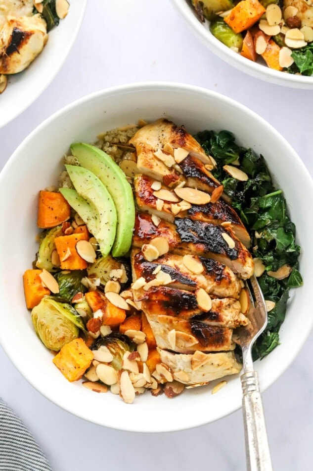 An overhead photo looking down at a chicken protein bowl with roasted veggies, slivered roasted almonds, avocado slices and sautéed kale. A fork rests inside the bowl.