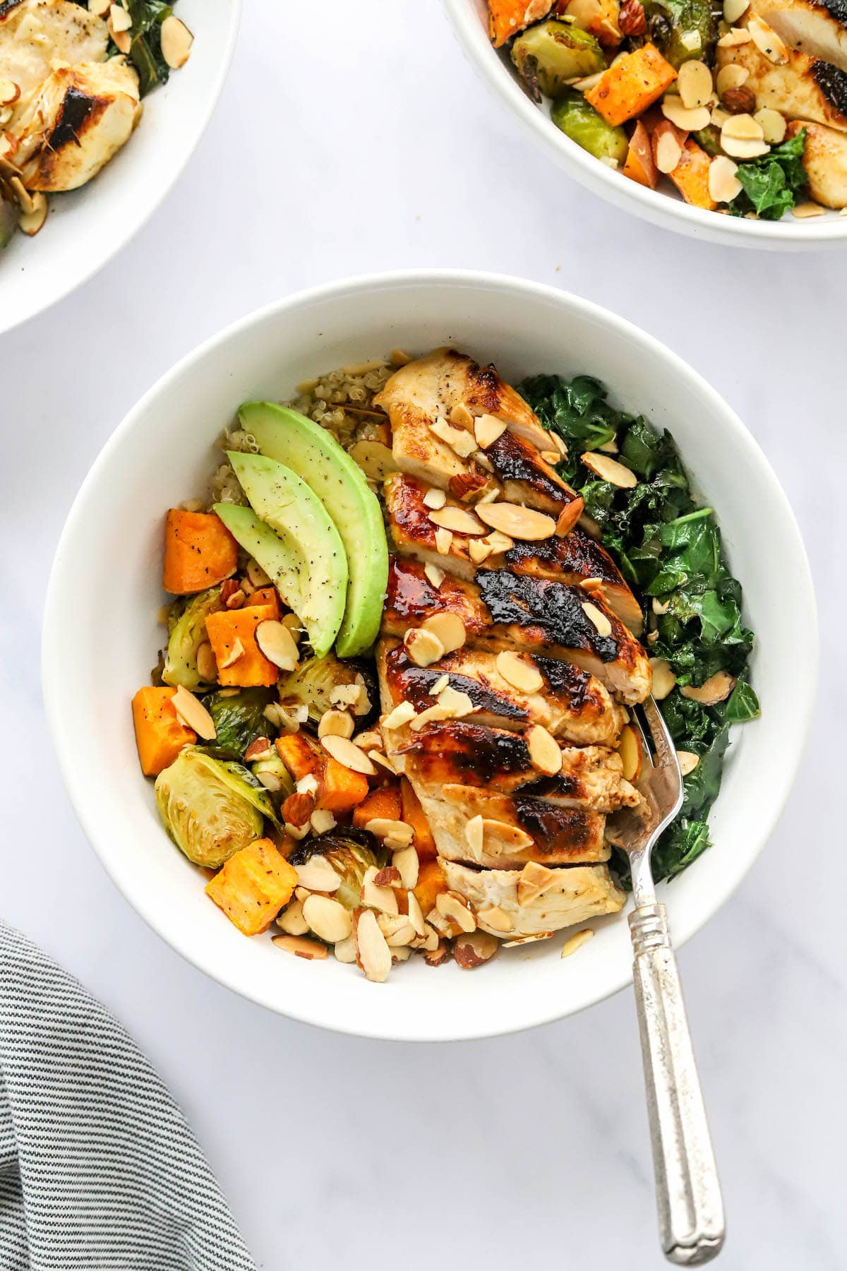 An overhead photo looking down at a chicken protein bowl with roasted veggies, slivered roasted almonds, avocado slices and sautéed kale. A fork rests inside the bowl.