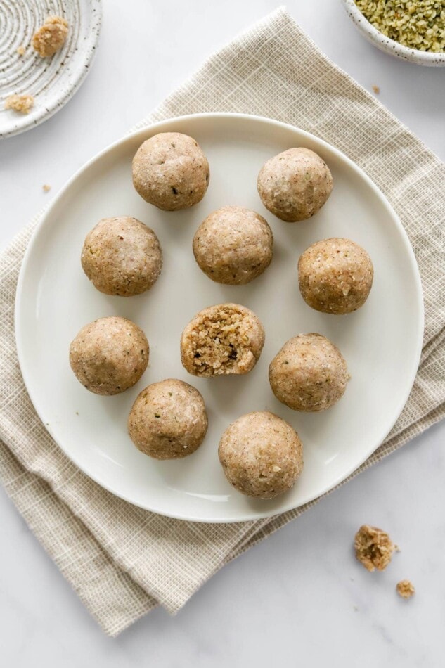 PIN THIS --> Vegan Coconut Protein Balls An overhead photo looking down on 10 coconut protein balls on a plate. The center ball has a bite taken out of it. The plate is on a khaki dish cloth.