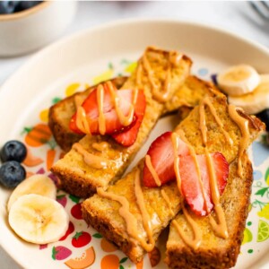 Slices of baby french toast on a plate topped with an almond butter drizzled and strawberry slices. There are banana and blueberry slices on the plate.