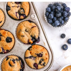 Blueberry muffins in a muffin tin. Next to the tin is a mug of coffee and bowl of blueberries.